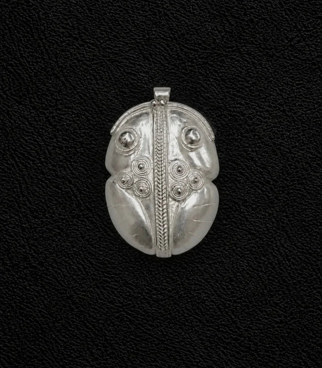 Un colgante plateado con intrincados grabados de estilo tribal, que incluyen una línea vertical central, detalles circulares y secciones en forma de hoja, sobre un fondo negro texturizado.