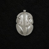 Un colgante plateado con intrincados grabados de estilo tribal, que incluyen una línea vertical central, detalles circulares y secciones en forma de hoja, sobre un fondo negro texturizado.