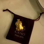 A gold pendant necklace displayed on a dark brown velvet drawstring pouch with the brand name CANO and 'Since 1970' printed on it, set against a light beige background.