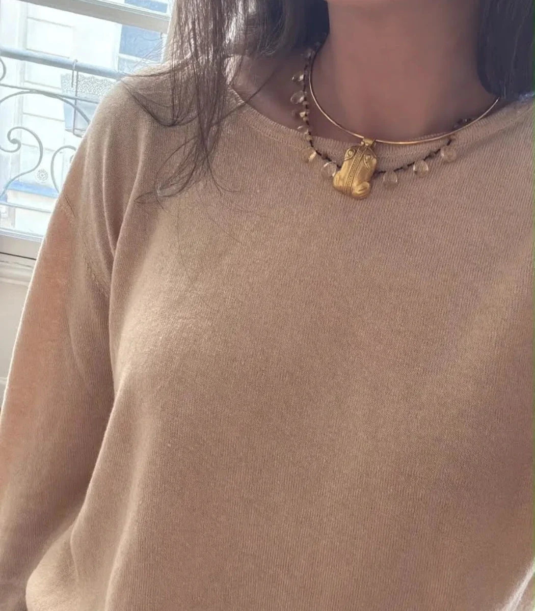 A close-up of a person wearing a beige knit sweater and a gold-toned beaded necklace with a small animal-shaped pendant, standing near a window.