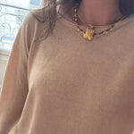 A close-up of a person wearing a beige knit sweater and a gold-toned beaded necklace with a small animal-shaped pendant, standing near a window.